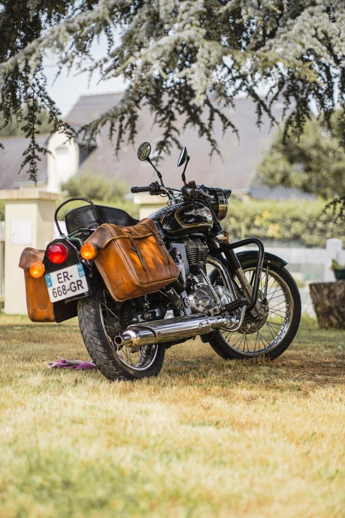 Vintage motorcycle with leather saddlebags parked on grass under tree shade on a sunny day.